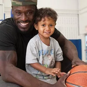 Man holding basketball and toddler son and smiling in YMCA indoor gymnasium