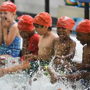 Group of kids with YMCA swim caps sitting on side of indoor pool and splashing water