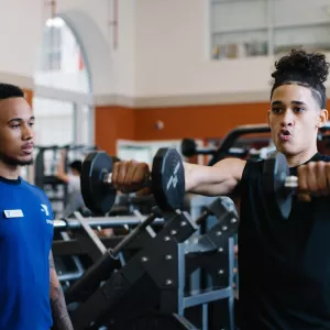 Fitness coach watches teen lifting dumbbells in YMCA weight lifting room at Ridgewood YMCA